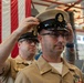 Two U.S. Navy Chief Petty Officers in khaki service uniforms are engaged in what appears to be a pinning or promotion ceremony. One sailor is placing or adjusting a Chief Petty Officer combination cover (cap) on the other sailor's head. A large American flag serves as a backdrop in what appears to be a hangar or large military facility.