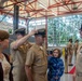 A military promotion or pinning ceremony is taking place inside what appears to be a naval facility or hangar. A service member is having a cover (hat) placed on his head by a fellow sailor, while a woman and a young girl observe alongside a senior officer. Additional uniformed personnel are visible in the background.