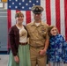 A U.S. Navy Chief Petty Officer in Service Khaki uniform poses with two family members — an adult woman and a young girl — in front of a large American flag. The setting appears to be a naval museum or ceremonial hall, with a ship or vessel visible in the background. The occasion appears to be a formal military ceremony, likely a promotion or retirement event.