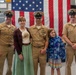 Three U.S. Navy personnel in tan service uniforms pose with a woman in civilian clothing and a young girl in front of a large American flag. The setting appears to be an indoor military facility or hangar, likely during a formal ceremony or official event. The group is smiling and appears to be posing for a commemorative photograph.