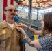 A military promotion or award ceremony is taking place indoors, likely at a naval facility. A woman with heavily tattooed arms is pinning a new insignia or medal onto the collar/chest area of a male sailor in a tan U.S. Navy uniform. An American flag and what appears to be a ship or vessel model are visible in the background.