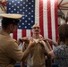 A military promotion or pinning ceremony is taking place in front of a large American flag. A senior officer in khaki uniform with a white cover and a civilian woman with tattoos are simultaneously pinning new rank insignia onto the collar of a service member in Navy khaki uniform. The setting appears to be an indoor military facility or hangar.