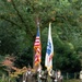 A U.S. Army color guard of four soldiers in dress uniforms marches across a well-maintained grass lawn, carrying the American flag and what appears to be a U.S. Army organizational flag. The ceremony appears to take place at a military cemetery or memorial garden, evidenced by white grave markers visible on the right side of the frame. The setting is lush and green with mature trees in the background.