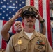 A U.S. Navy Chief Petty Officer is having his combination cover (cap) placed on his head during what appears to be a Chief Petty Officer pinning or advancement ceremony. A civilian or retired military individual stands behind him, carefully positioning the cover. The ceremony takes place in front of a large American flag, consistent with a formal military advancement ritual.
