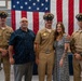 Five individuals pose for a group photo in front of a large American flag in what appears to be a military facility or naval vessel hangar. Three individuals are wearing U.S. Navy Chief Petty Officer dress uniforms with covers (hats), while one civilian man and one civilian woman stand among them. The setting and attire suggest a military ceremony, likely a promotion or pinning event.
