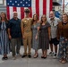A group of seven people pose for a formal photograph in front of a large American flag inside what appears to be a military facility or hangar. One individual is wearing a U.S. Navy dress khaki uniform with a cover (officer's cap), suggesting this is a military ceremony or promotion event. The remaining six individuals are dressed in civilian attire, likely family members or guests.
