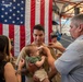 A U.S. Navy promotion or pinning ceremony is taking place inside what appears to be a military facility or hangar. A young sailor in khaki uniform is holding an infant while an older civilian man pins a new insignia or rank device onto his collar. Family members are present, and a large American flag serves as the backdrop.