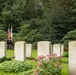 A military cemetery ceremony is taking place among rows of Commonwealth-style war graves with rounded headstones bearing carved emblems and crosses. A color guard carrying what appears to be the American flag and at least one other flag stands between the grave rows in the background. The setting is lush and green with dense trees and flowering plants surrounding the memorial site.