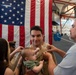 A U.S. Navy service member in tan/khaki uniform is being pinned with new insignia by two individuals — a woman on his left and an older man on his right — in what appears to be a promotion ceremony. The sailor is holding a baby while smiling, with a large American flag displayed prominently in the background. The setting appears to be an indoor military facility or hangar.