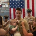 A U.S. Navy promotion or pinning ceremony is taking place in front of a large American flag. A sailor in khaki uniform is holding an infant while two women pin insignia or rank devices onto his collar or chest. Two senior enlisted Navy personnel in covers (hats) stand in the background observing the ceremony.