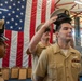 A U.S. Navy promotion or pinning ceremony is taking place in what appears to be a hangar or gymnasium facility. A service member is having a Chief Petty Officer cover (hat) placed on his head by another sailor, while a female officer stands at attention to the left. An American flag serves as the backdrop, and award cases are visible on a table in the background.