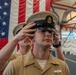 A U.S. Navy sailor is being fitted with a Chief Petty Officer combination cover (cap) during what appears to be a CPO pinning or advancement ceremony. A large American flag is prominently displayed in the background, indicating a formal military ceremony setting. A second sailor in the same tan uniform is visible in the background.