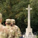 Military personnel in camouflage uniforms are gathered at what appears to be a memorial ceremony before a large stone Cross of Sacrifice, a design commonly associated with Commonwealth War Graves Commission cemeteries. The cross features a bronze sword affixed to its face, which is a hallmark of Sir Reginald Blomfield's iconic WWI memorial design. Empty wreath-laying stands are visible in front of the monument, suggesting a formal commemorative event.