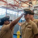 A U.S. Navy ceremony is taking place inside what appears to be a naval facility or gymnasium. A female Navy chief petty officer is adjusting or placing a combination cover (hat) on a male sailor who is wearing ribbons and warfare devices on his khaki uniform. An American flag and Navy recruitment banners are visible in the background.
