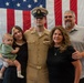 A U.S. Navy servicemember in dress khaki uniform poses with what appears to be his family in front of a large American flag. The group consists of five individuals including a young woman holding an infant, the uniformed sailor, a middle-aged woman, and an older man. The setting appears to be an official military ceremony or promotion event.