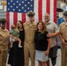 A group of seven people pose for a photo in front of a large American flag in what appears to be a military facility or hangar. Three individuals are wearing U.S. Navy uniforms, including two in Service Khaki and one in Service Dress Khaki with a combination cover, suggesting a military ceremony or promotion event. The group includes civilians, one of whom is holding an infant, indicating this is likely a family celebration.