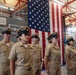A formal U.S. Navy ceremony is taking place inside what appears to be a naval hangar or facility. Several Navy personnel in khaki service uniforms and combination covers are standing at attention in formation before a large American flag. A speaker or official can be seen at a podium to the right side of the image.
