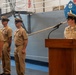 A U.S. Navy chief petty officer is delivering a speech at a podium during what appears to be a formal military ceremony, likely a chief petty officer pinning or advancement ceremony. Several other Navy personnel in khaki service uniforms stand in formation to the left. The event appears to be held inside a naval facility or museum, with a ship's hull and naval insignia visible in the background.