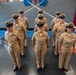 A group of United States Navy personnel in khaki service uniforms (Service Uniform Charlie/Delta) are standing in formation inside what appears to be a naval facility or ship hangar bay. The personnel include both male and female sailors wearing combination covers (peaked caps) with chief petty officer insignia and various service ribbons. The setting suggests a formal ceremony or official event.