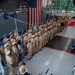 A formal U.S. Navy military ceremony is taking place inside what appears to be a ship's hangar bay or large indoor military facility. A large formation of Navy personnel in khaki service uniforms stands at attention while a small brass ensemble performs in the foreground. A large American flag is prominently displayed on the back wall, and a blue military unit guidon flag is visible within the formation.