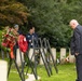 A solemn memorial ceremony is taking place at what appears to be a Commonwealth war cemetery, identifiable by the uniform white headstones. A man in a dark suit with medals is bowing his head in respect before several wreath stands holding poppy wreaths and floral arrangements. Two individuals in blue clothing stand in the background observing the ceremony.