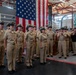 A group of U.S. Navy personnel in khaki service uniforms are gathered in what appears to be a hangar or large indoor military facility for a formal ceremony. A large American flag is prominently displayed in the background. The personnel appear to be reacting positively, with some laughing and applauding, suggesting a celebratory or recognition event.