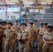 A group of U.S. Navy personnel in khaki service uniforms are gathered in what appears to be a training facility or naval museum space. The group appears to be in a celebratory or informal moment, with several members laughing and smiling. One central figure is holding a small stuffed animal toy, and an American flag is prominently displayed in the background.