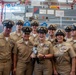 A group of approximately nine U.S. Navy personnel in khaki service uniforms are posing together for a group photo at what appears to be an official military ceremony or event. One person in the center is holding a small stuffed animal toy resembling a dog or bear. The setting appears to be inside a naval facility decorated with signal flags, a U.S. flag, and a ship or vessel structure in the background.