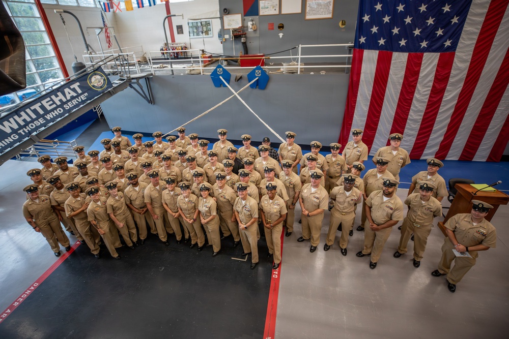 A large group of U.S. Navy personnel in tan/khaki service uniforms are assembled for a formal group photograph inside what appears to be a naval training facility or gymnasium. An oversized American flag is prominently displayed on the right wall, and a banner reading 'WHITEHAT' and 'THOSE WHO SERVE' is visible on the left. The setting suggests a graduation, ceremony, or official unit photo event.