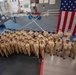 A large group of U.S. Navy personnel in tan/khaki service uniforms are assembled for a formal group photograph inside what appears to be a naval training facility or gymnasium. An oversized American flag is prominently displayed on the right wall, and a banner reading 'WHITEHAT' and 'THOSE WHO SERVE' is visible on the left. The setting suggests a graduation, ceremony, or official unit photo event.