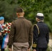 A military commemoration ceremony is taking place at what appears to be a war cemetery, with uniformed personnel walking toward a wreath-laying site. Two central figures in military dress uniforms are seen from behind approaching a man holding a floral wreath with red, white, and blue colors. Stone grave markers are visible in the background, consistent with a military burial ground.