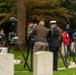 A military memorial ceremony is taking place at what appears to be a military cemetery, with uniformed personnel placing floral wreaths on stands among white grave markers. Two central figures — one in U.S. Army olive drab uniform and one in a dark naval-style uniform with white cap — are placing wreaths, while additional military and civilian participants stand in the background. Multiple national flags including the U.S. flag are visible, suggesting a multinational commemorative event.