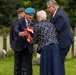 A solemn memorial ceremony is taking place in what appears to be a military cemetery, identifiable by the uniform white headstones in the background. Four individuals — including two men in military berets, one elderly woman, and one man in a suit — are gathered around a red poppy wreath. The scene strongly suggests a remembrance or commemoration event, likely associated with Commonwealth war graves.