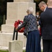 A commemorative wreath-laying ceremony is taking place at what appears to be a military or war cemetery. An elderly woman in a patterned jacket is placing a red poppy wreath on a stand in front of a large stone memorial cross, assisted by a man in a dark suit. Several other attendees are visible in the background among white headstones.