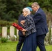 An elderly woman carrying a red poppy wreath is being escorted by a man in a navy suit through what appears to be a military cemetery. The scene suggests a commemorative ceremony, likely a Remembrance Day or war memorial service. Rows of white headstones are visible in the background, consistent with Commonwealth War Graves Commission cemeteries.