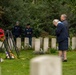 A solemn memorial ceremony is taking place at a military cemetery, likely a Commonwealth War Graves Commission site, identifiable by the uniform white headstones. Two individuals in the foreground bow their heads in respect before poppy wreaths on stands, while uniformed personnel and civilians observe in the background. The setting and red poppy wreaths are consistent with Remembrance Day or a similar commemorative event.
