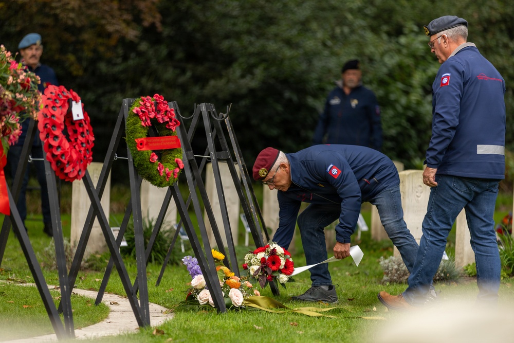 A memorial ceremony is taking place at what appears to be a military cemetery, with individuals wearing matching navy blue jackets and maroon/red berets laying flowers at grave markers. Multiple wreaths made of red poppies are displayed on black stands, suggesting a commemorative event such as a WWII remembrance ceremony. The patches visible on the jackets appear to reference the 82nd Airborne Division insignia.