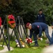 A memorial ceremony is taking place at what appears to be a military cemetery, with individuals wearing matching navy blue jackets and maroon/red berets laying flowers at grave markers. Multiple wreaths made of red poppies are displayed on black stands, suggesting a commemorative event such as a WWII remembrance ceremony. The patches visible on the jackets appear to reference the 82nd Airborne Division insignia.