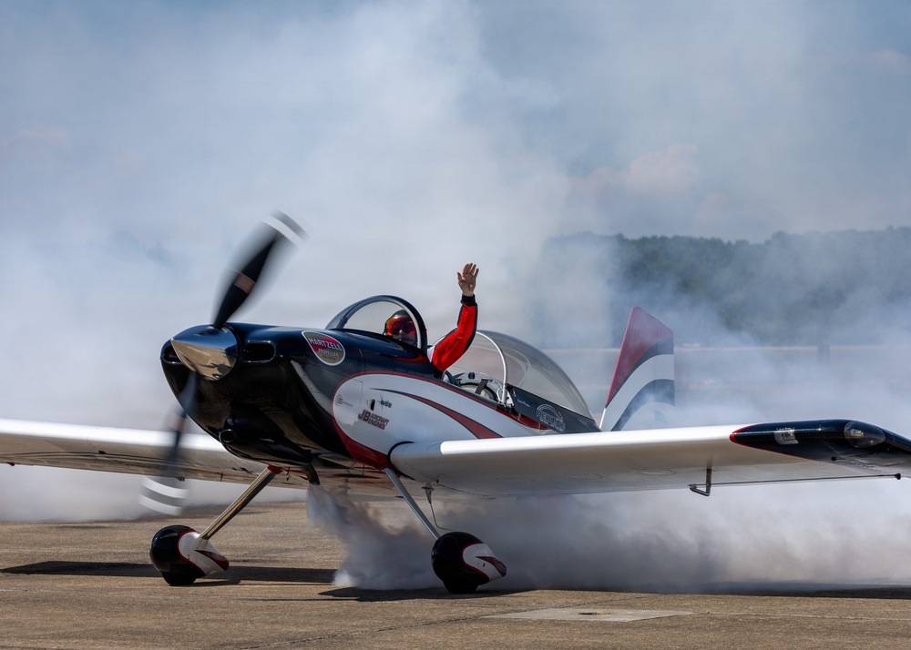 A pilot in a red flight suit and helmet waves from the open cockpit of a small aerobatic aircraft on a tarmac. The aircraft, decorated in a black, white, and red livery with visible sponsor logos including 'Hartzell', is surrounded by dense white smoke, suggesting a smoke system is active. This appears to be an airshow performance or post-performance taxi.