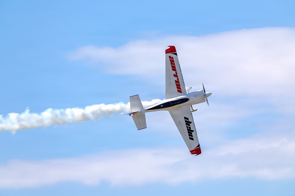 A single-engine propeller aircraft is performing an aerobatic maneuver, appearing to be in an inverted or steeply banked roll. The aircraft is white with red and black markings, trailing a white smoke stream behind it. The image was taken from below against a partly cloudy blue sky.