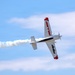 A single-engine propeller aircraft is performing an aerobatic maneuver, appearing to be in an inverted or steeply banked roll. The aircraft is white with red and black markings, trailing a white smoke stream behind it. The image was taken from below against a partly cloudy blue sky.