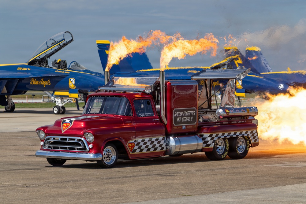 A jet-powered custom truck named 'Hayden Proffitt's Hot Streak II' performs a fire-breathing demonstration on an airshow tarmac. The vehicle, styled after a classic 1957 Chevrolet pickup in candy red with checkered accents, shoots massive flames from its jet engines. In the background, multiple U.S. Navy Blue Angels F/A-18 Super Hornet aircraft are parked on the flight line.