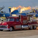 A jet-powered custom truck named 'Hayden Proffitt's Hot Streak II' performs a fire-breathing demonstration on an airshow tarmac. The vehicle, styled after a classic 1957 Chevrolet pickup in candy red with checkered accents, shoots massive flames from its jet engines. In the background, multiple U.S. Navy Blue Angels F/A-18 Super Hornet aircraft are parked on the flight line.