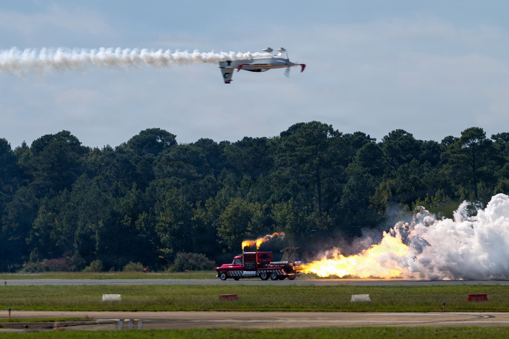 An airshow performance featuring two simultaneous acts: an inverted aircraft trailing white smoke in the upper portion of the frame, and a jet-powered truck on the runway below producing large flames and white smoke clouds. The event appears to take place at a military or civilian airfield with a tree line visible in the background. The combination of the aerial and ground performances suggests a coordinated airshow demonstration.