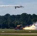 An airshow performance featuring two simultaneous acts: an inverted aircraft trailing white smoke in the upper portion of the frame, and a jet-powered truck on the runway below producing large flames and white smoke clouds. The event appears to take place at a military or civilian airfield with a tree line visible in the background. The combination of the aerial and ground performances suggests a coordinated airshow demonstration.