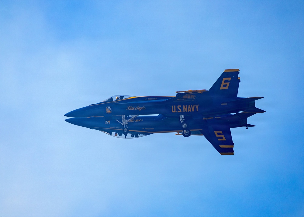 Two U.S. Navy Blue Angels F/A-18 Hornet jets are captured in extremely close formation flight against a blue sky. The aircraft are numbered 5 and 6, flying in an inverted mirror formation with their undersides nearly touching. The image showcases the precision flying characteristic of the Blue Angels flight demonstration squadron.