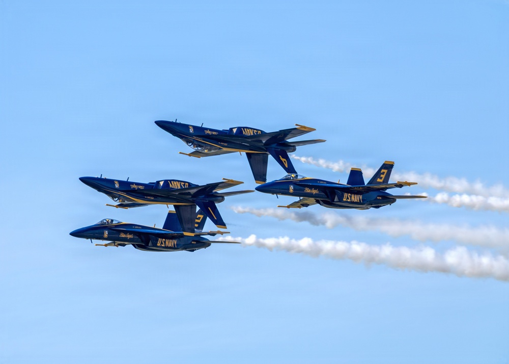 Four U.S. Navy Blue Angels aircraft are performing a tight formation flight maneuver against a clear blue sky. The jets, painted in their iconic blue and gold livery, are flying in a diamond-like stacked formation while producing white contrails. The aircraft are identifiable as F/A-18 Hornets or Super Hornets bearing visible unit numbers.