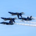 Four U.S. Navy Blue Angels aircraft are performing a tight formation flight maneuver against a clear blue sky. The jets, painted in their iconic blue and gold livery, are flying in a diamond-like stacked formation while producing white contrails. The aircraft are identifiable as F/A-18 Hornets or Super Hornets bearing visible unit numbers.