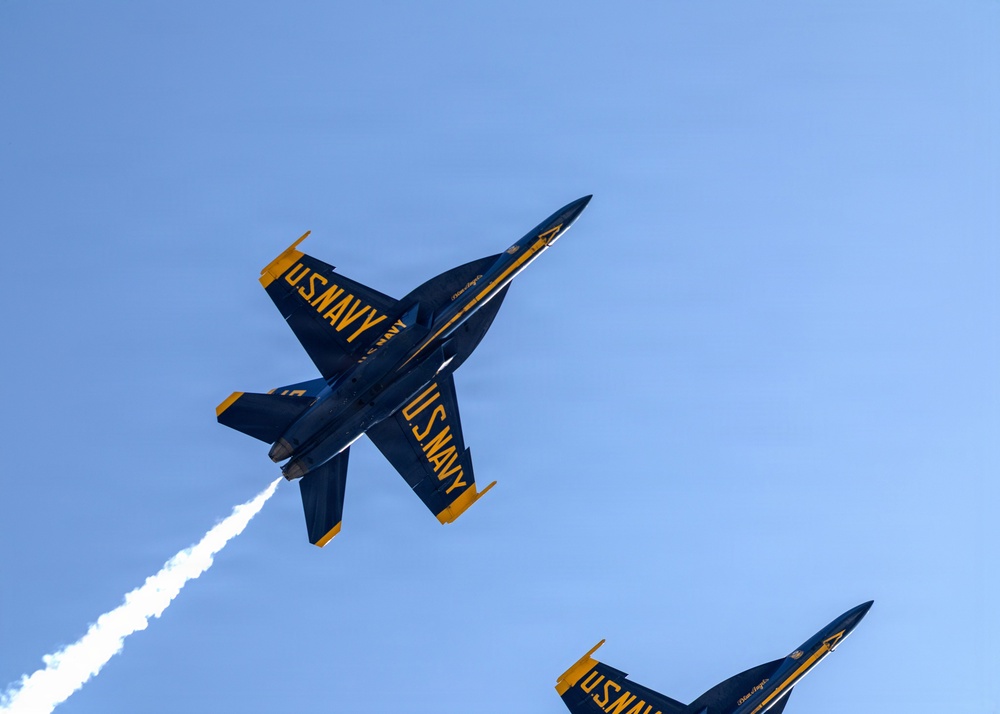 Two U.S. Navy Blue Angels F/A-18 Hornet jets are captured in a close formation maneuver during an airshow performance. The aircraft are photographed from below against a clear blue sky, with one jet emitting a white smoke trail. The distinctive blue and gold livery with 'U.S. NAVY' lettering is clearly visible on both aircraft.
