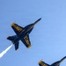 Two U.S. Navy Blue Angels F/A-18 Hornet jets are captured in a close formation maneuver during an airshow performance. The aircraft are photographed from below against a clear blue sky, with one jet emitting a white smoke trail. The distinctive blue and gold livery with 'U.S. NAVY' lettering is clearly visible on both aircraft.